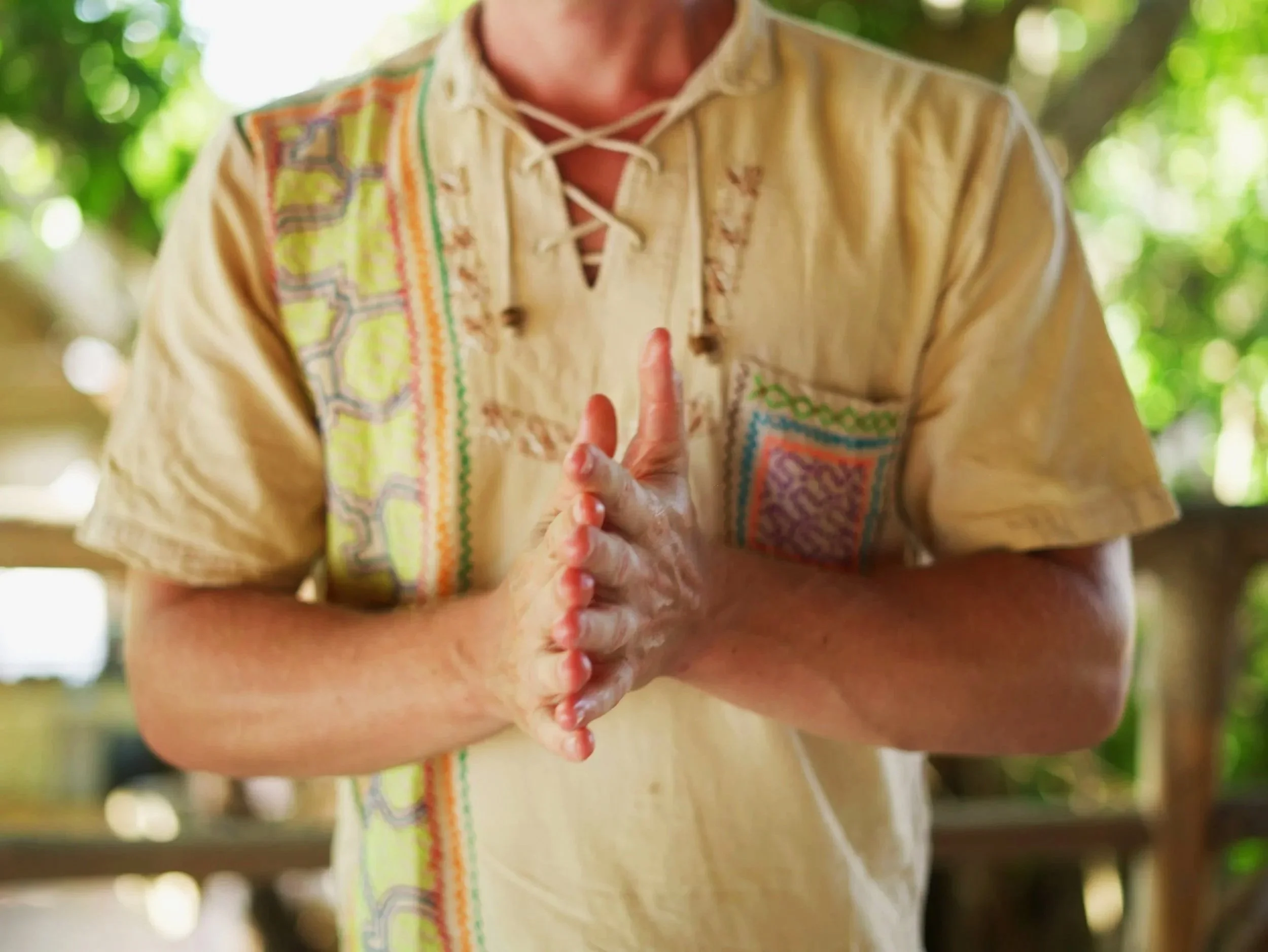 Der Shipibo Masseur Michael Heitmann trägt ein beiges Hemd mit bunten Shipibo Mustern und hält die Hände in Gebetshaltung in die Kamera. Die Fingerspitzen sind so ausgerichtet, dass sie übereinander stehen und wie eine Energiekette aussehen.