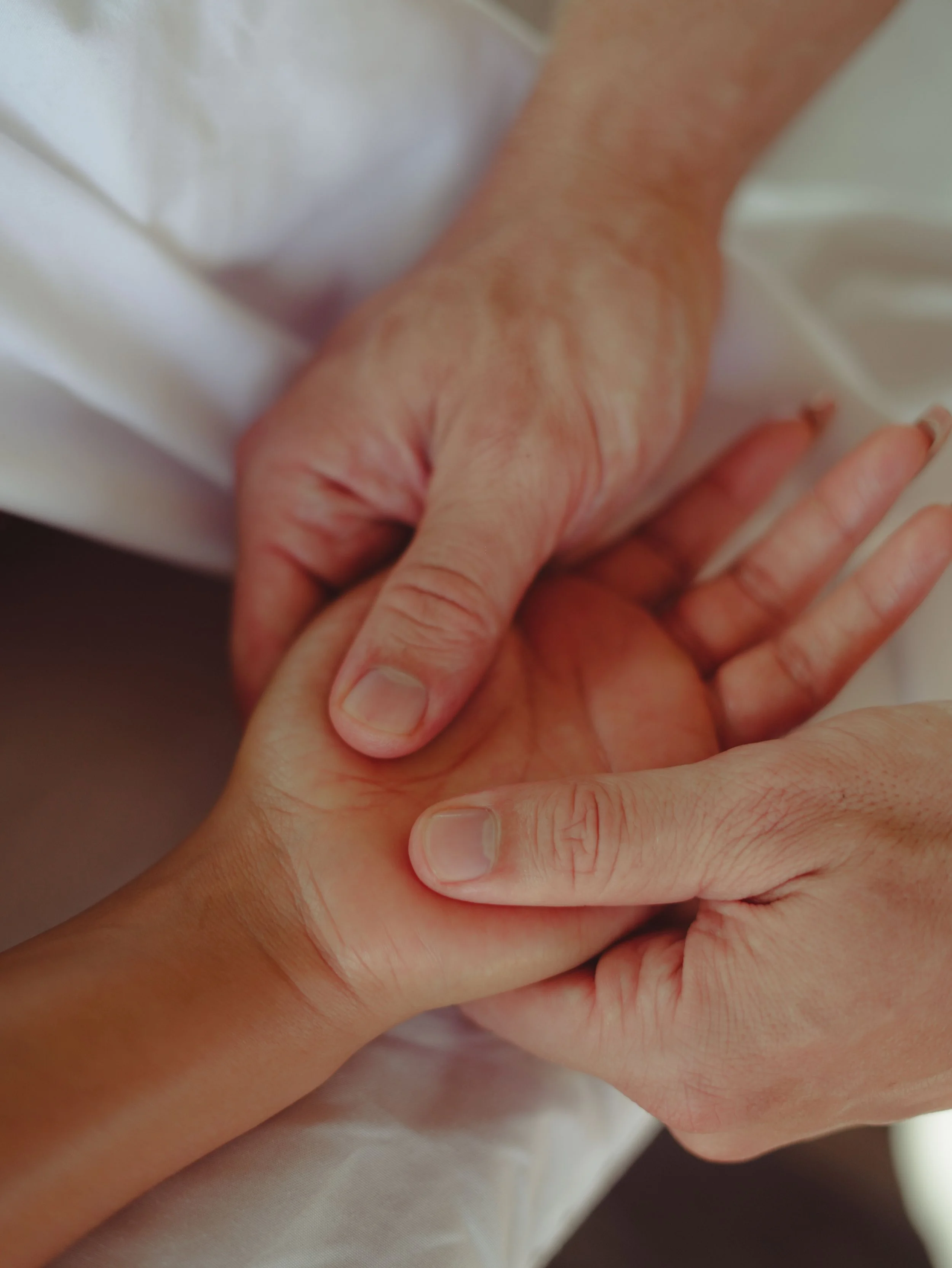 Nahaufnahme einer Handmassage mit Massageöl. Die nach oben zeigende Hand einer Frau wird von außen von beiden Händen des Massagegebers umfasst, während die Daumen des Masseurs die Handwurzel kneten.