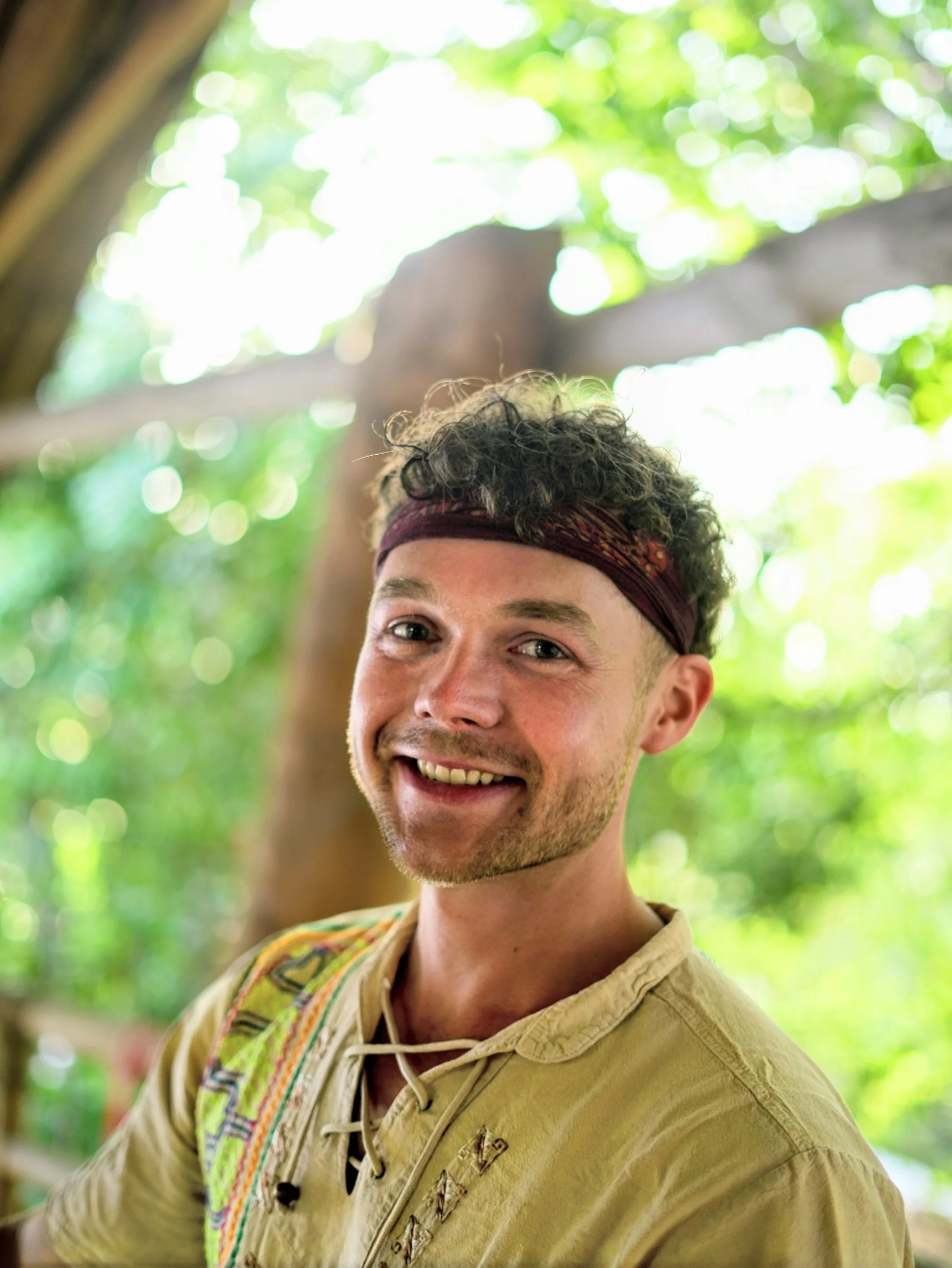 Ein junger Mann mit lockigem Haar, rotem Stirnband und traditioneller Shipibo Zeremoniebekleidung, lächelnd im Freien bei sonnigem Wetter mit grünen Blättern im Hintergrund. Es ist Michael Heitmann, Der Inhaber von Shipibo Massage Osnabrück.