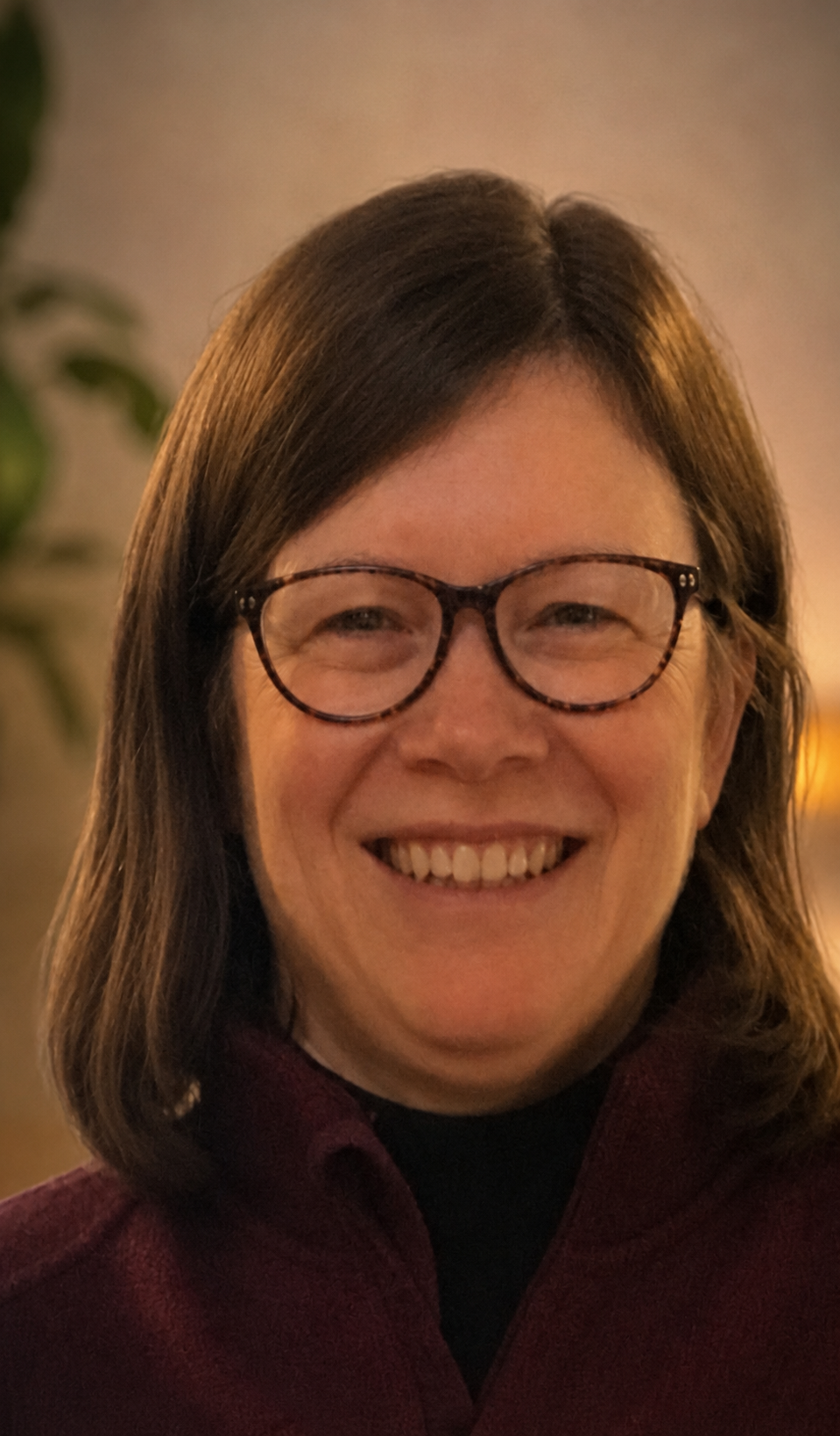 A woman with shoulder-length brown hair, glasses, and a big smile, wearing a dark red or maroon top, in an indoor setting.
