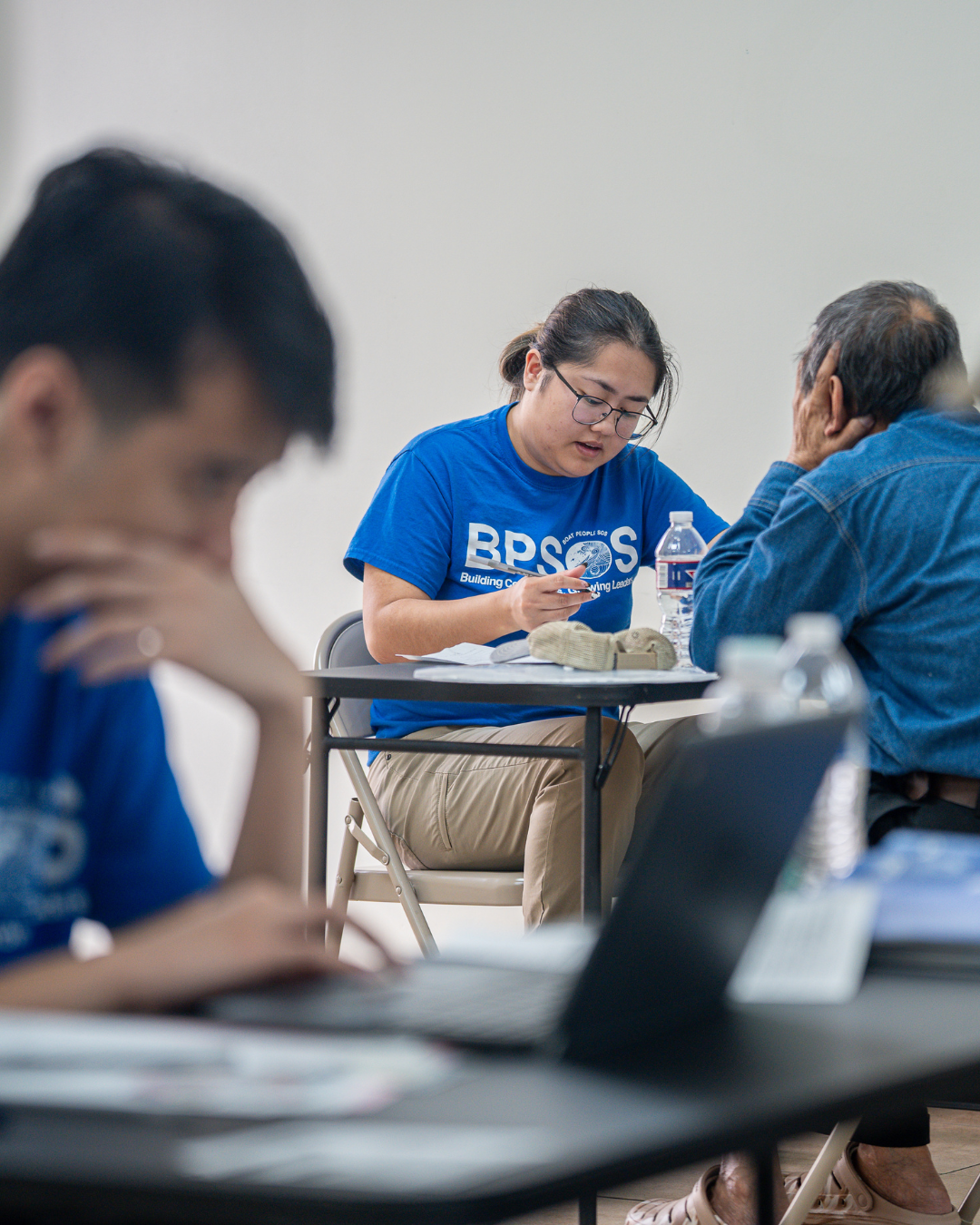 A woman studying at a table with a water bottle and papers, wearing a blue t-shirt with 'BPSOS' printed on it, in a classroom setting with other students around her.