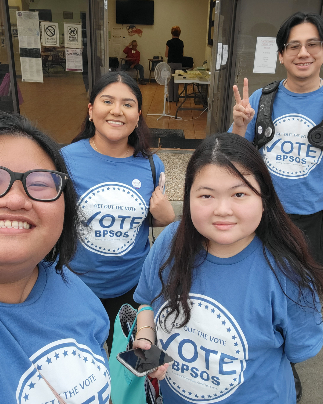 Four people wearing blue 'Vote BPSOS' T-shirts, standing outside a building, smiling at the camera. One person is making a peace sign, another holds a phone, and they seem to be participating in a voting or community event.