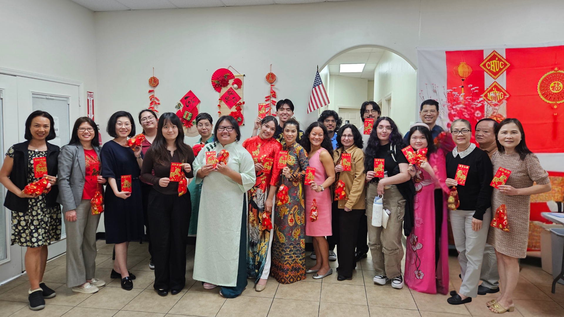 A group of people gathered in a room celebrating Lunar New Year, holding red envelopes and decorations. The room is decorated with red banners, lanterns, and an American flag is visible in the background.