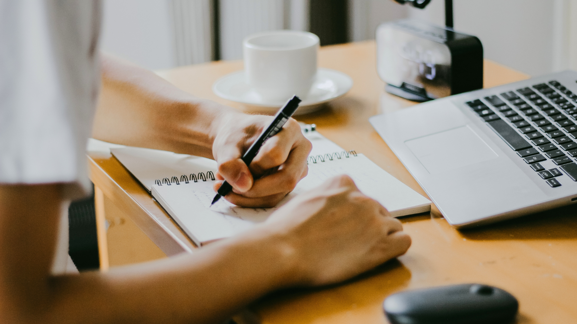 A person sitting at a wooden desk, writing in a spiral notebook with a black pen. On the desk, there is a silver laptop, a cup of coffee on a saucer, and a small black and white paper puncher.