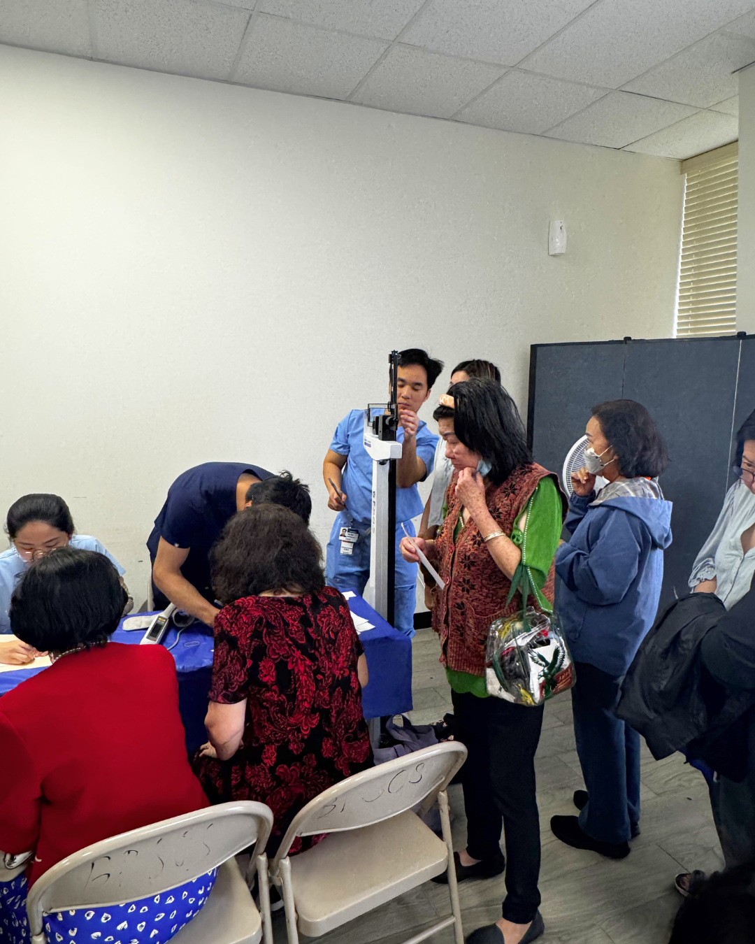 Group of people at a health screening or registration desk, with medical staff and a height measurement scale, in a room with plain white walls and window blinds.