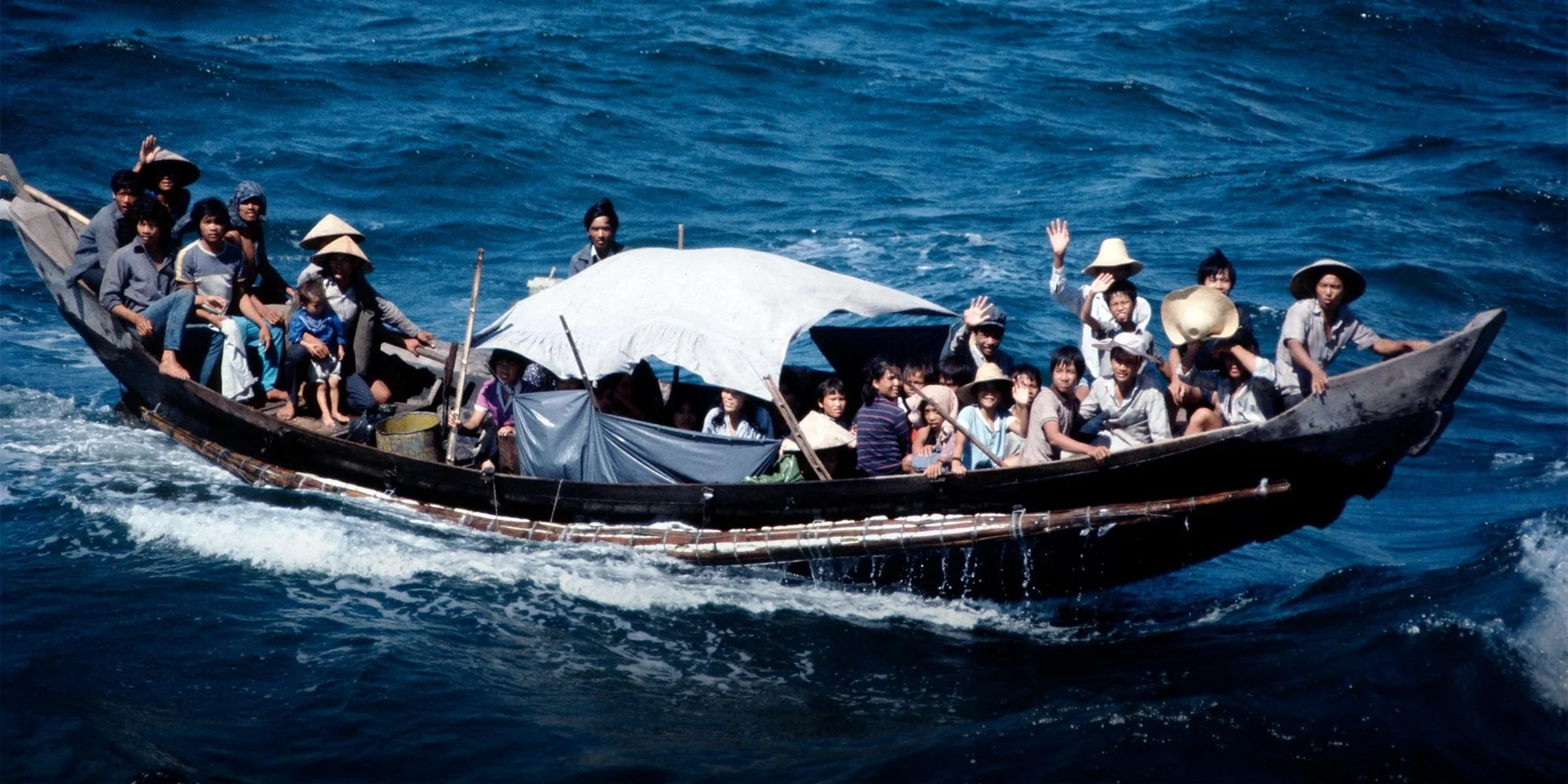 A boat filled with numerous people, many wearing traditional conical hats, navigating through the ocean with blue water surrounding it.