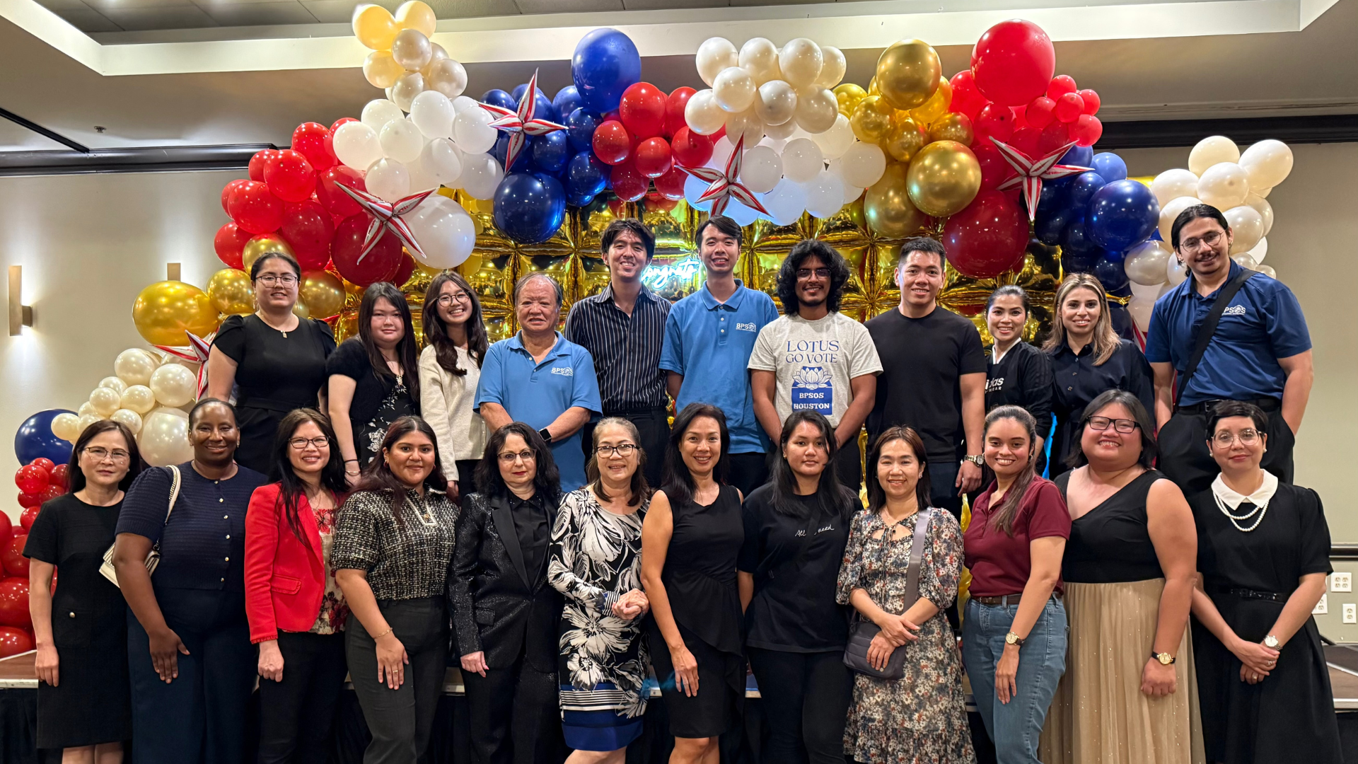 Group of diverse people at a celebration event with balloon decorations in red, white, blue, gold, and star-shaped balloons in the background.