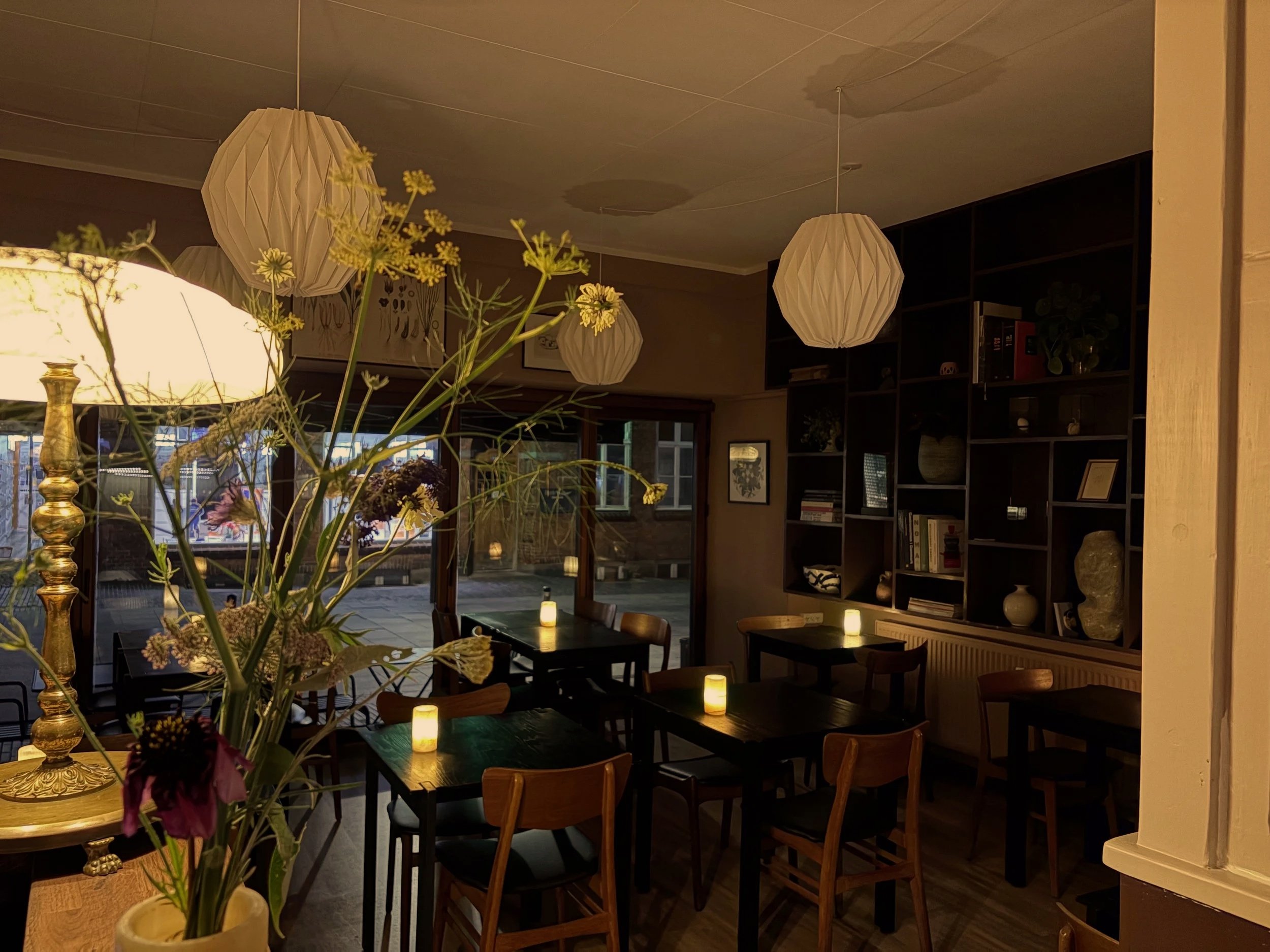 Indendørs restaurant med borddekorationer, blomster, og hængende paplampeskærme, oplyst af små stearinlys på bordene.