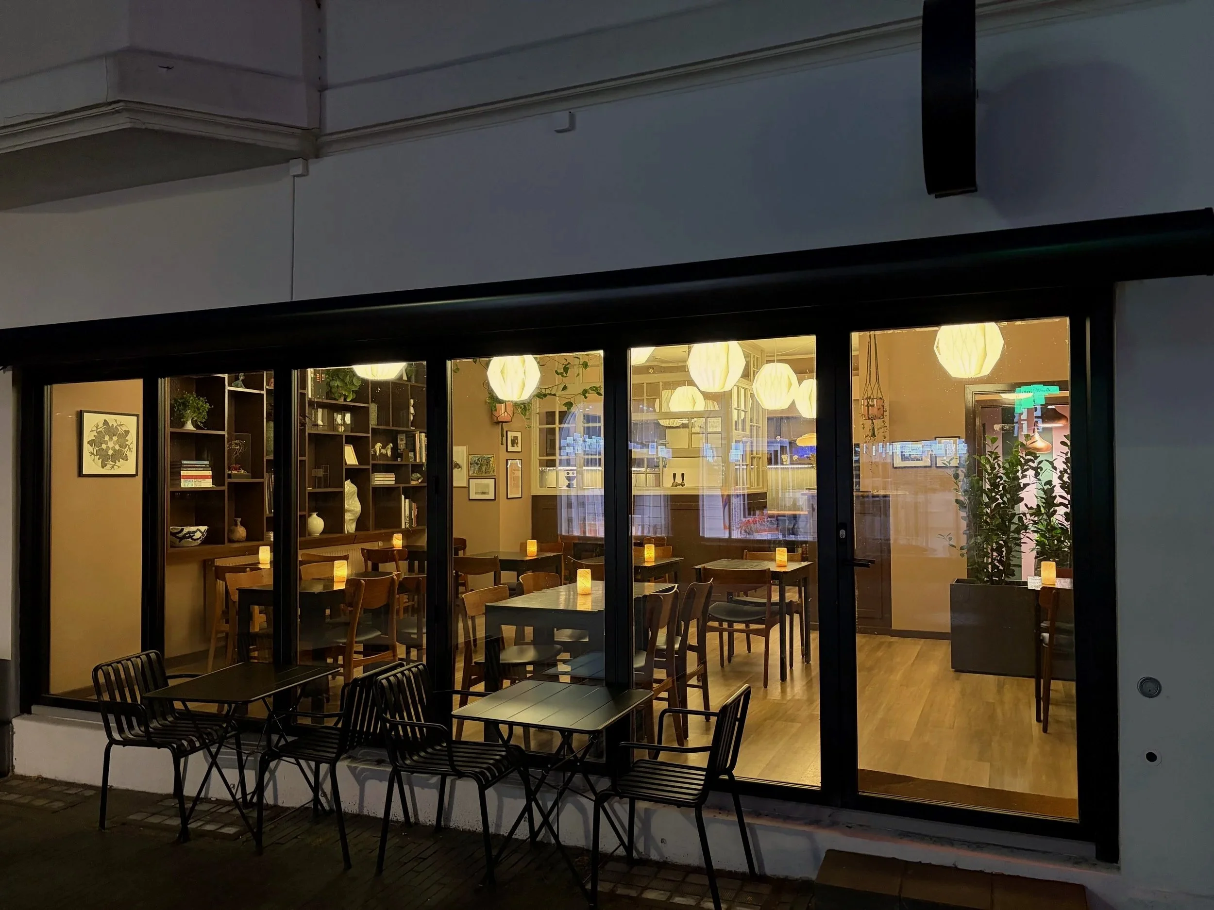 Indendørs cafe med spiseborde, bløde belysning, bøger og dekorative planter set gennem store glasporte.