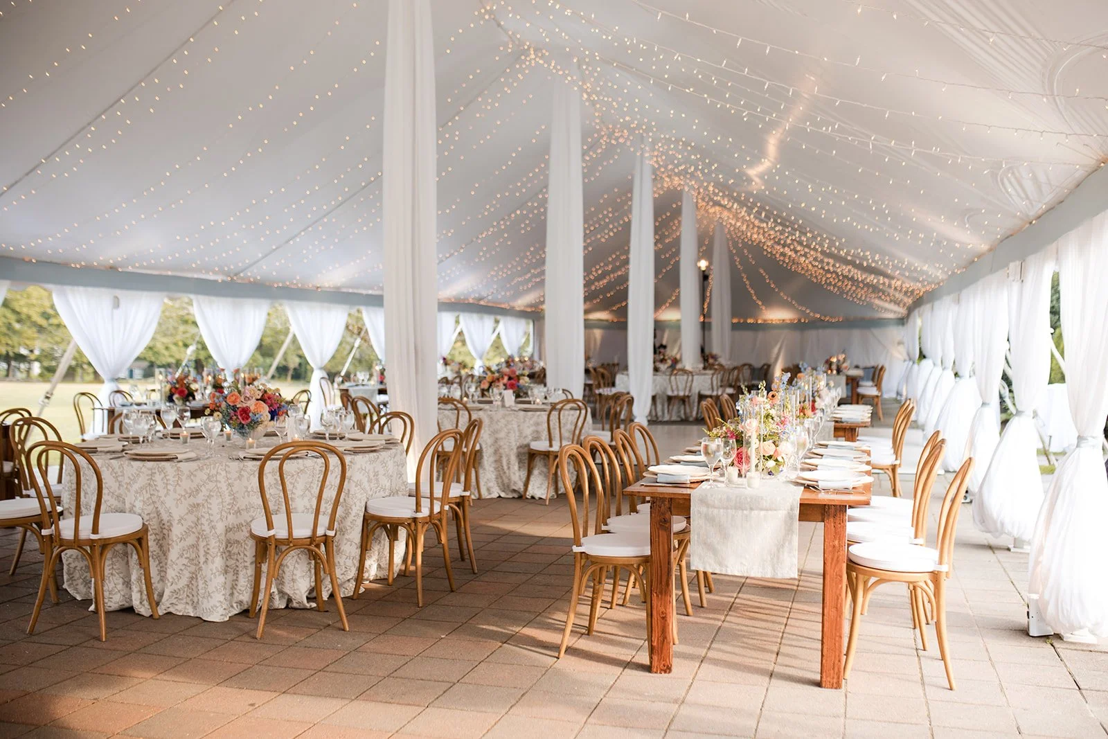 Elegant outdoor wedding reception under a white tent with hanging string lights, decorated tables with floral centerpieces, candles, and white drapery in the background.