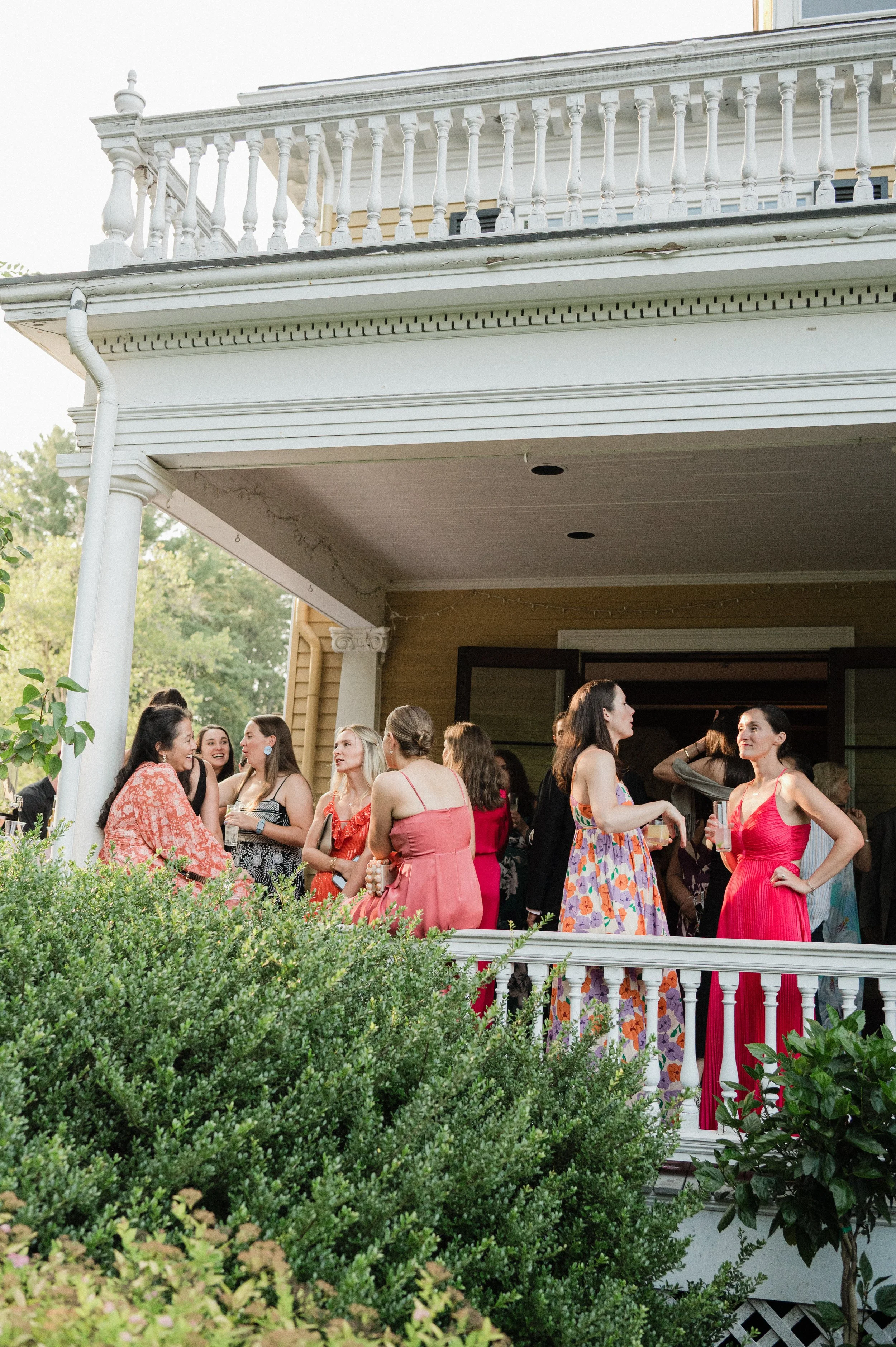 Group of women gathered on a house's porch, dressed for a party or gathering, with some holding drinks, engaging in conversation.