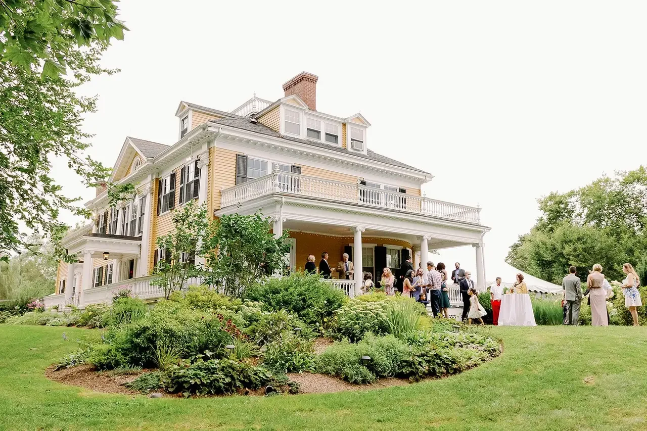 People gathered on the porch and lawn of a large, historic yellow house with white trim, celebrating outdoors on a garden wedding or party day.