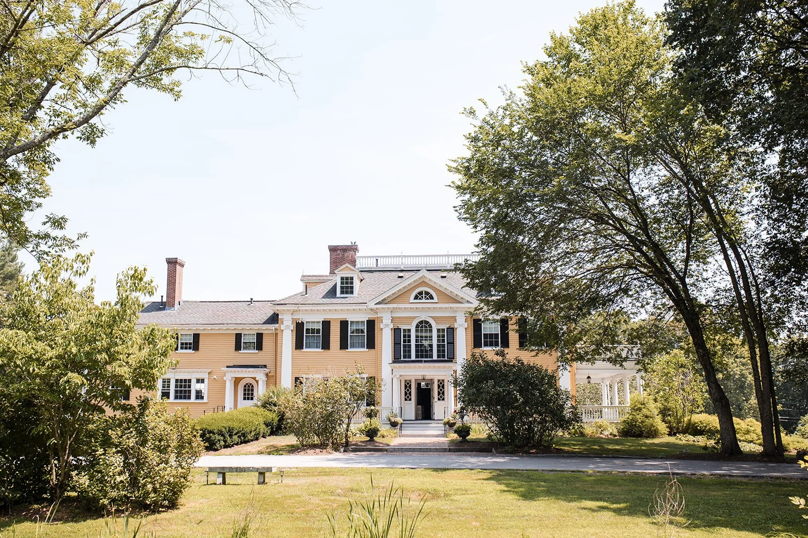 A large yellow house with white columns and black shutters, surrounded by green trees and shrubs, under a clear sky.