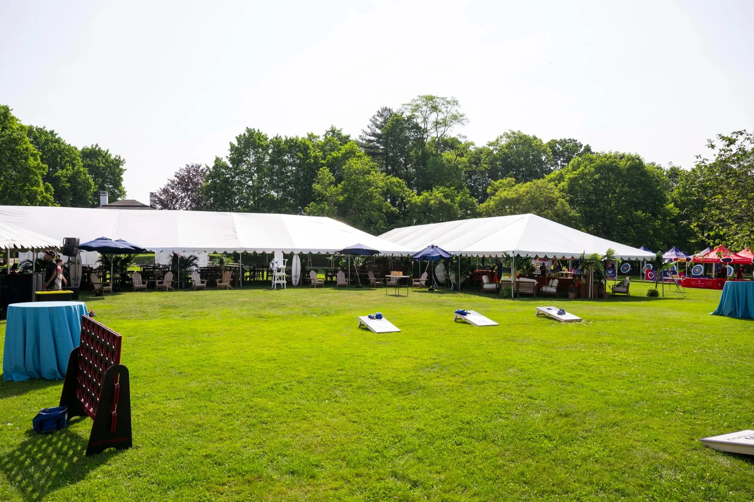 An outdoor event setup with white tents, tables, chairs, and decorations on a grassy field, surrounded by tall green trees and a clear sky.