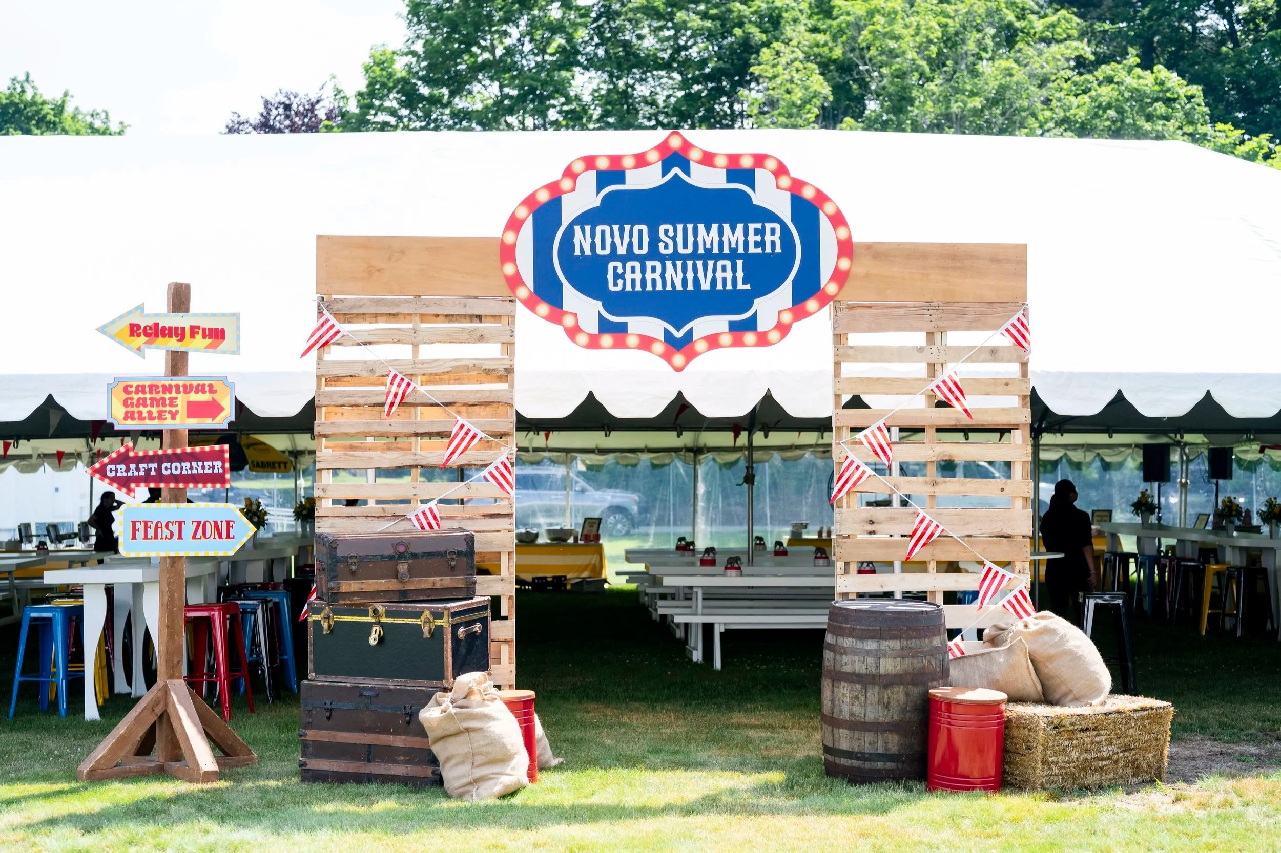 Outdoor carnival setup with a large sign reading "Novo Summer Carnival" and various directional signs pointing to relay fun, carnival game alley, craft corner, and feast zone. Decorated with striped bunting, surrounded by tables, barrels, and sacks u