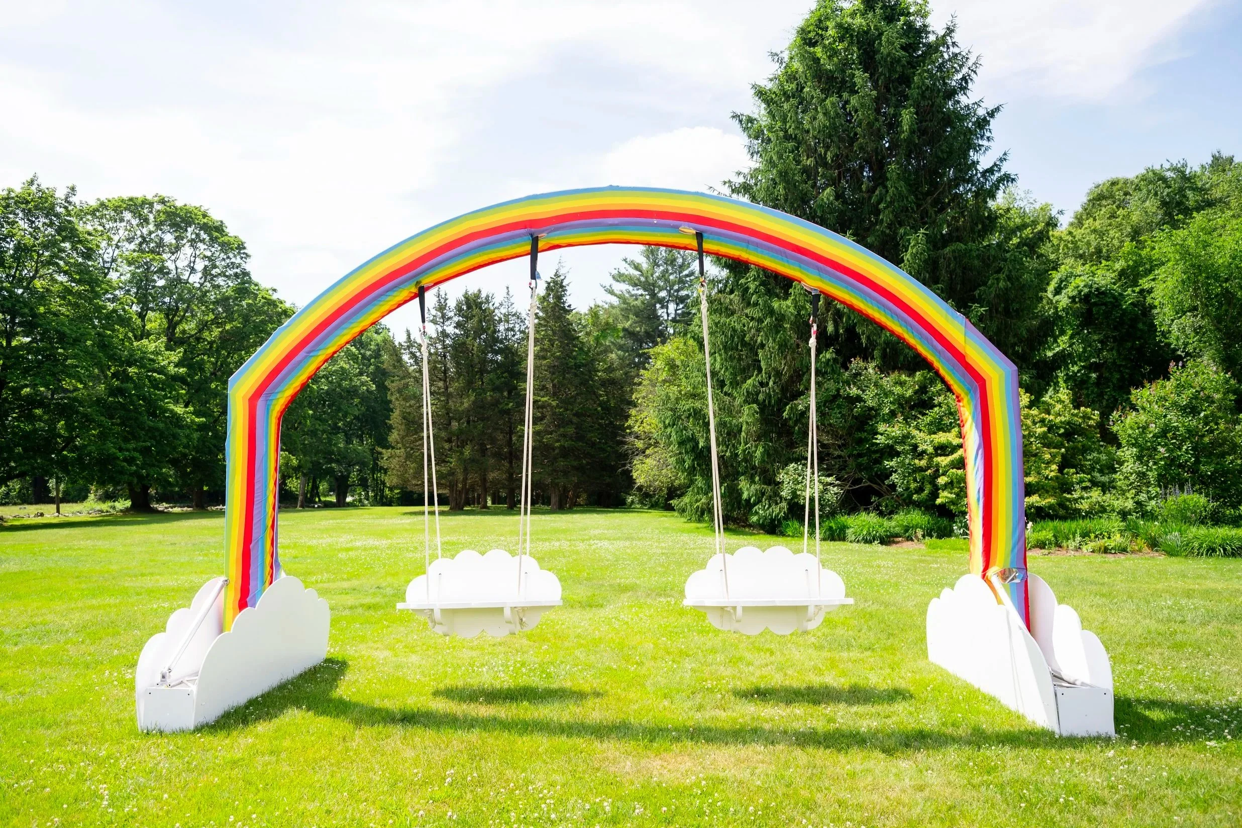 A rainbow-colored swing set shaped like a rainbow arch with white cloud bases, set on a grassy field with trees in the background.