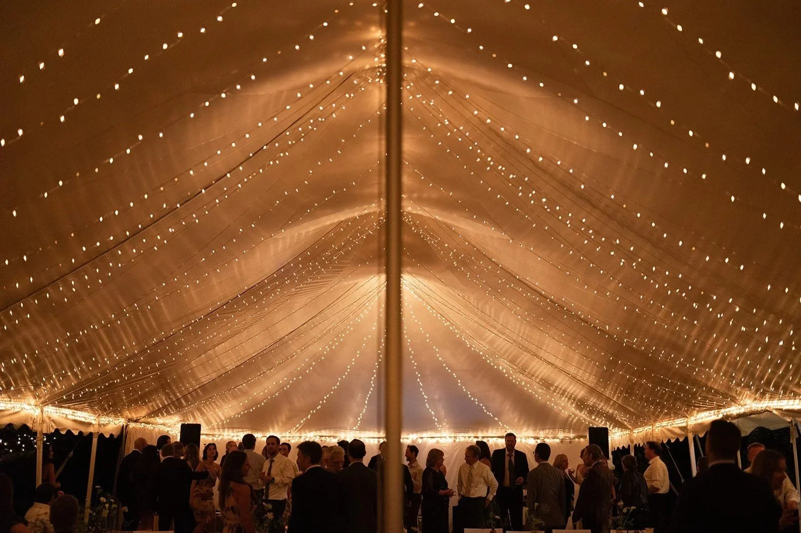 People gathered and dancing under a large tent decorated with string lights at an evening event or party.