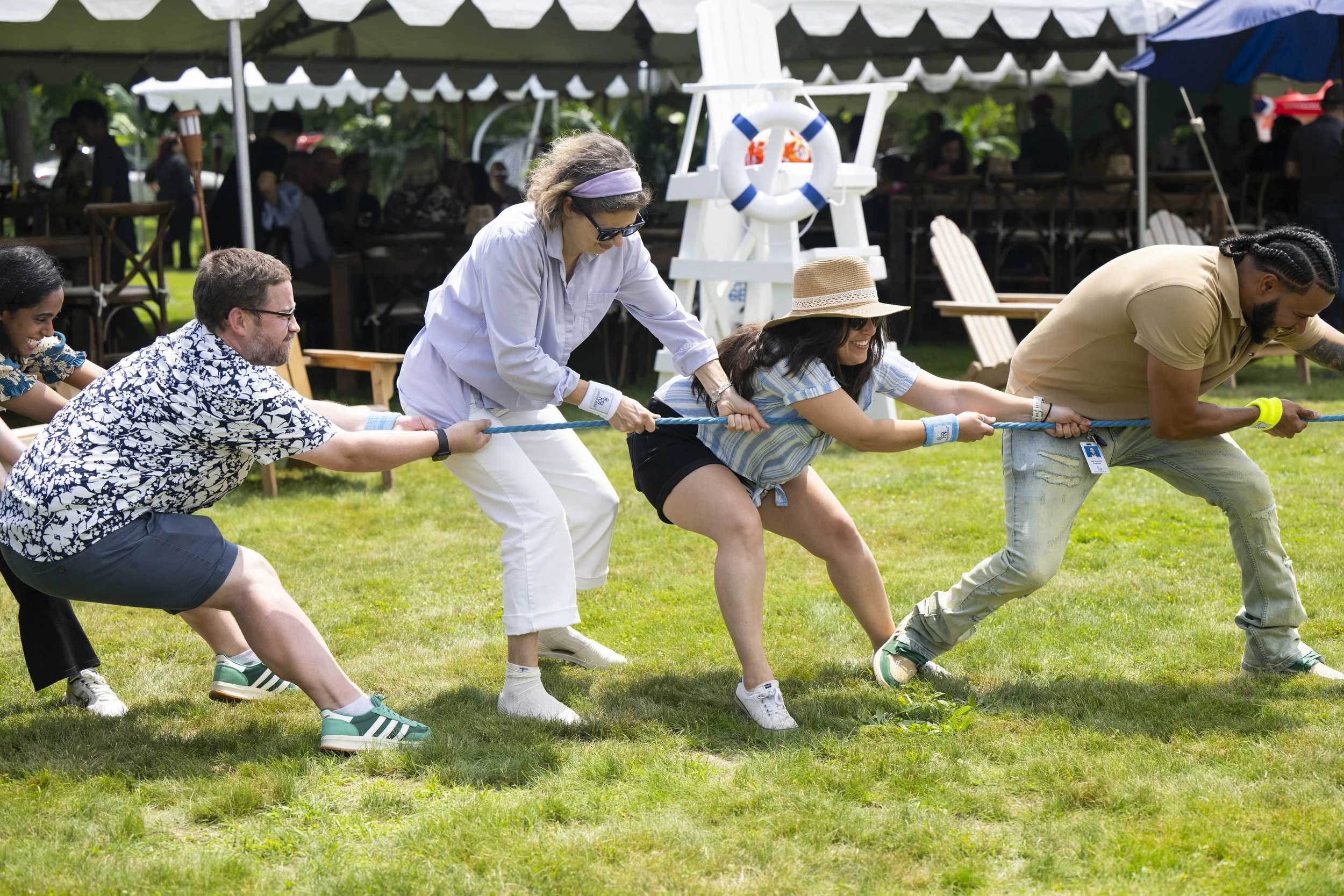 People participating in a tug-of-war game outdoors at a gathering or party, with a tent, tables, and chairs in the background.