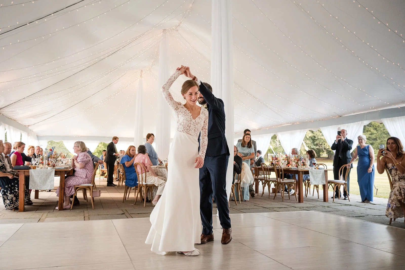 A bride and groom sharing a dance at their wedding reception under a decorated tent with string lights, surrounded by seated guests and onlookers taking photos.