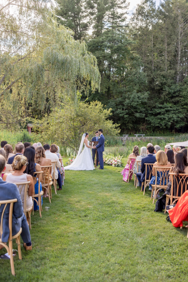 A wedding ceremony with a bride and groom exchanging vows outdoors in a lush green park surrounded by trees, with guests seated on either side.