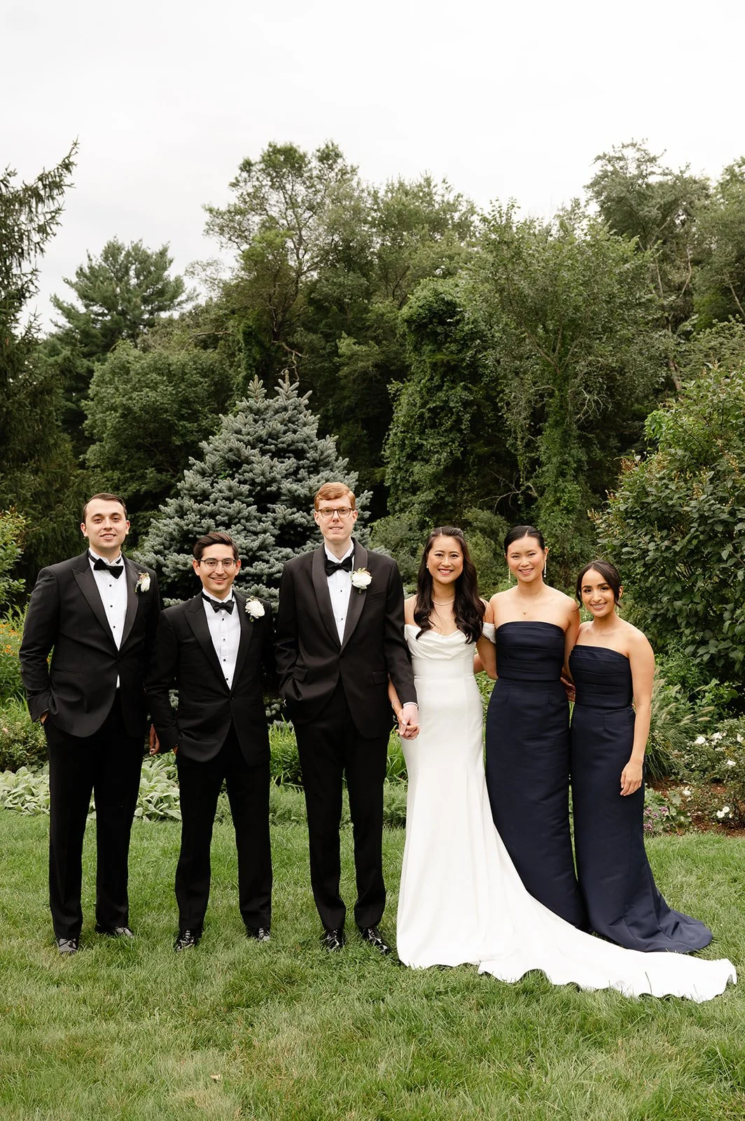 A group of six diverse people standing outdoors on grass during a wedding, with a background of trees and green foliage. They are dressed in formal attire, including tuxedos and bridesmaid dresses.