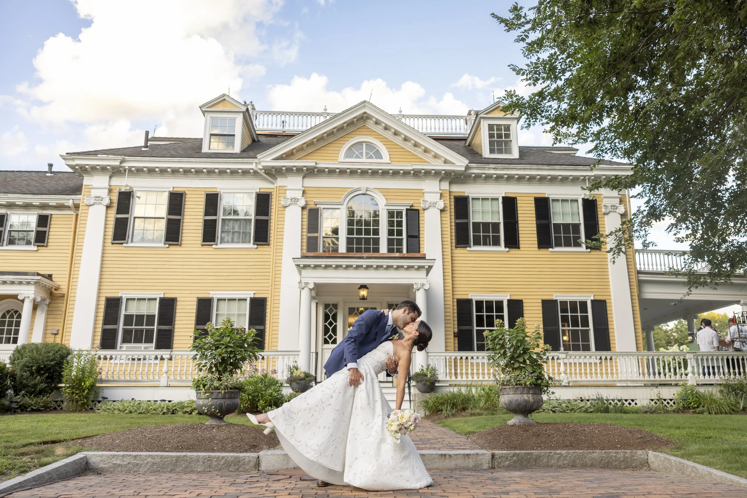A newlywed couple sharing a kiss in front of a large yellow house with white trim, black shutters, and a garden. The groom dips the bride, who is holding a bouquet of flowers.