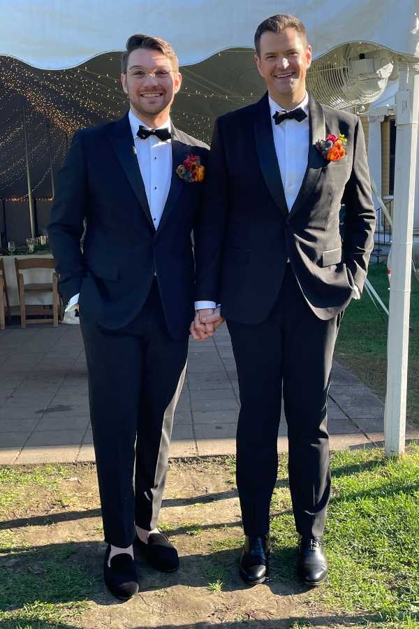 Two men in tuxedos holding hands, standing outdoors at a wedding reception under a white tent with string lights, smiling at the camera.