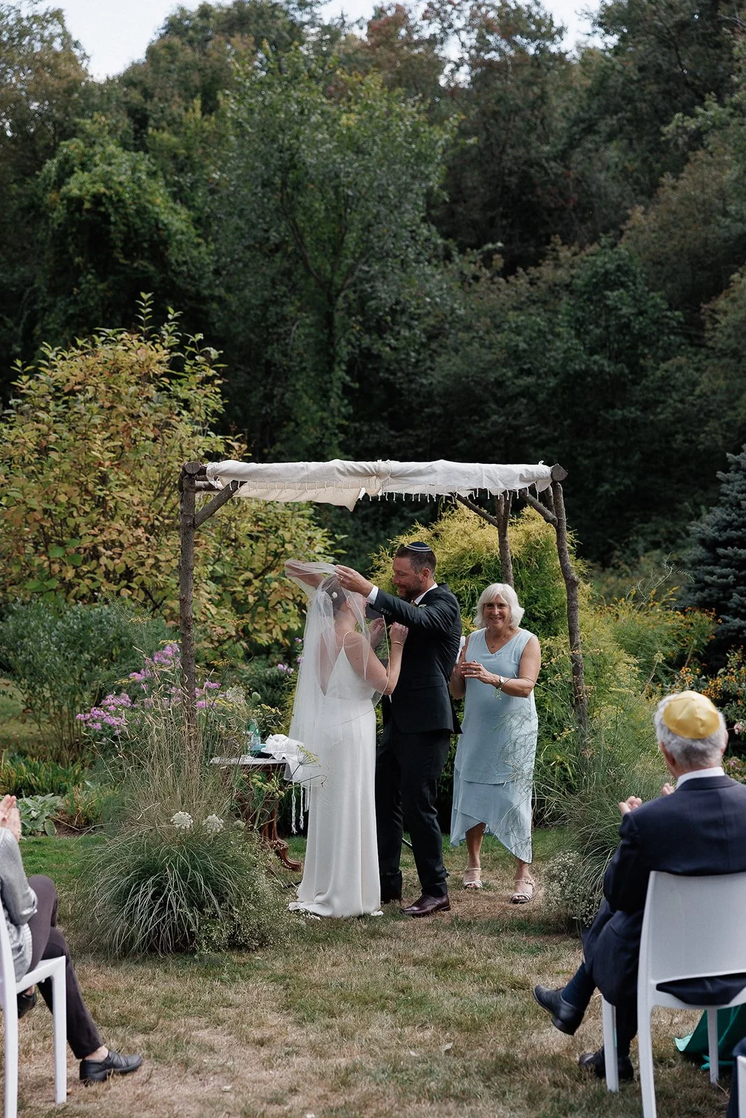 A couple getting married outdoors under a wooden canopy with white fabric. The groom is lifting the bride's veil. An officiant and guests are present, with some guests clapping and wearing traditional head coverings. The setting is a lush garden with colorful plants and trees.