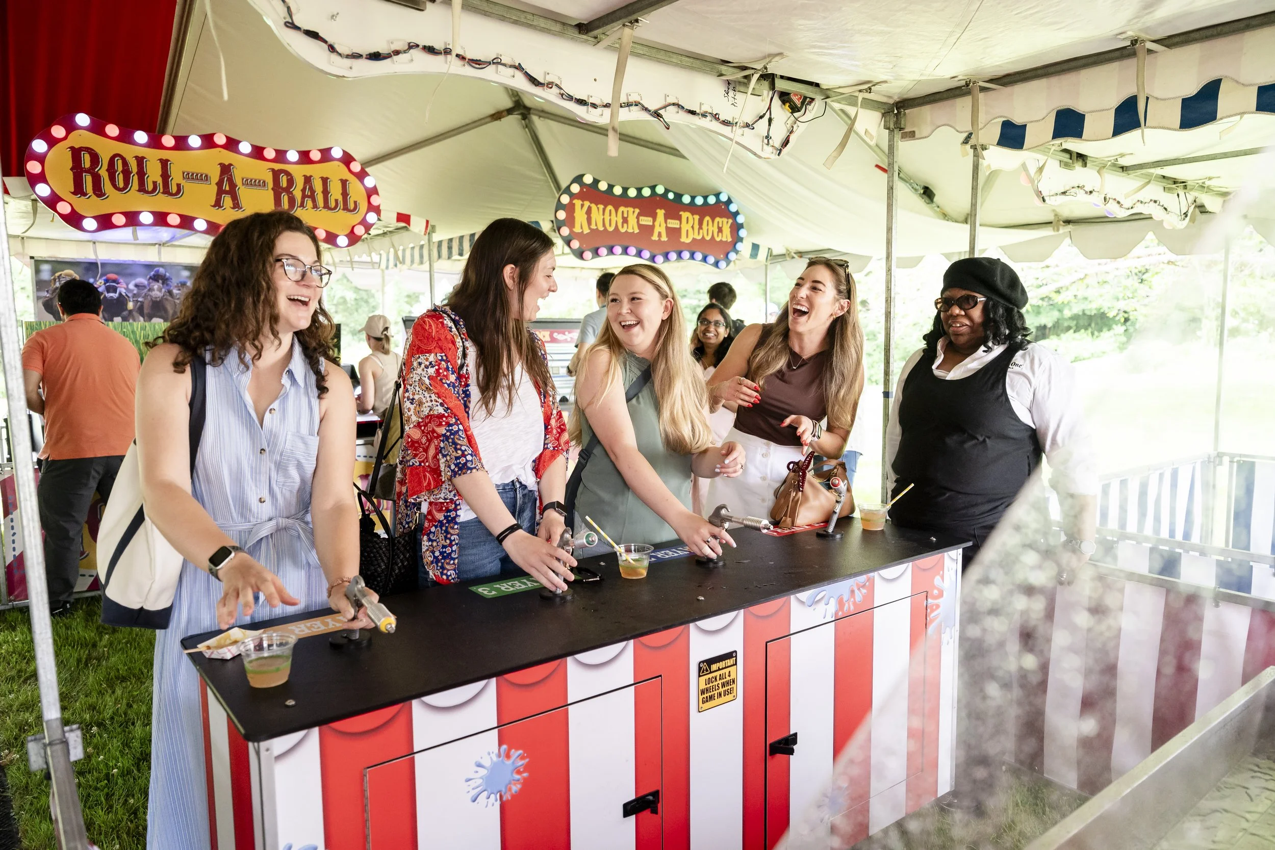 A group of four women enjoying a carnival game booth under a tent, with colorful signs reading 'Roll-A-Ball' and 'Knock-A-Block'. They are smiling and holding mallets. There are other people and trees in the background.