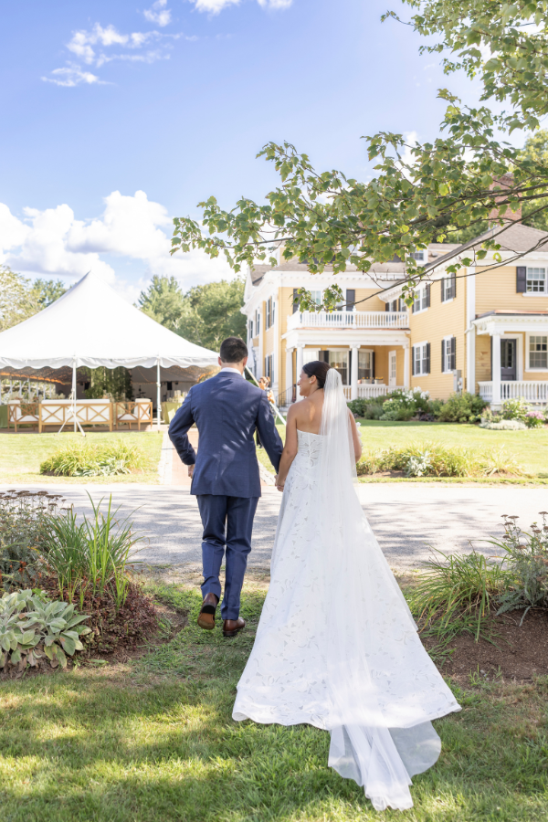 Bride and groom walking hand in hand toward a yellow house with a white porch, outdoor wedding setup with a white tent, green lawn, and trees under a blue sky.