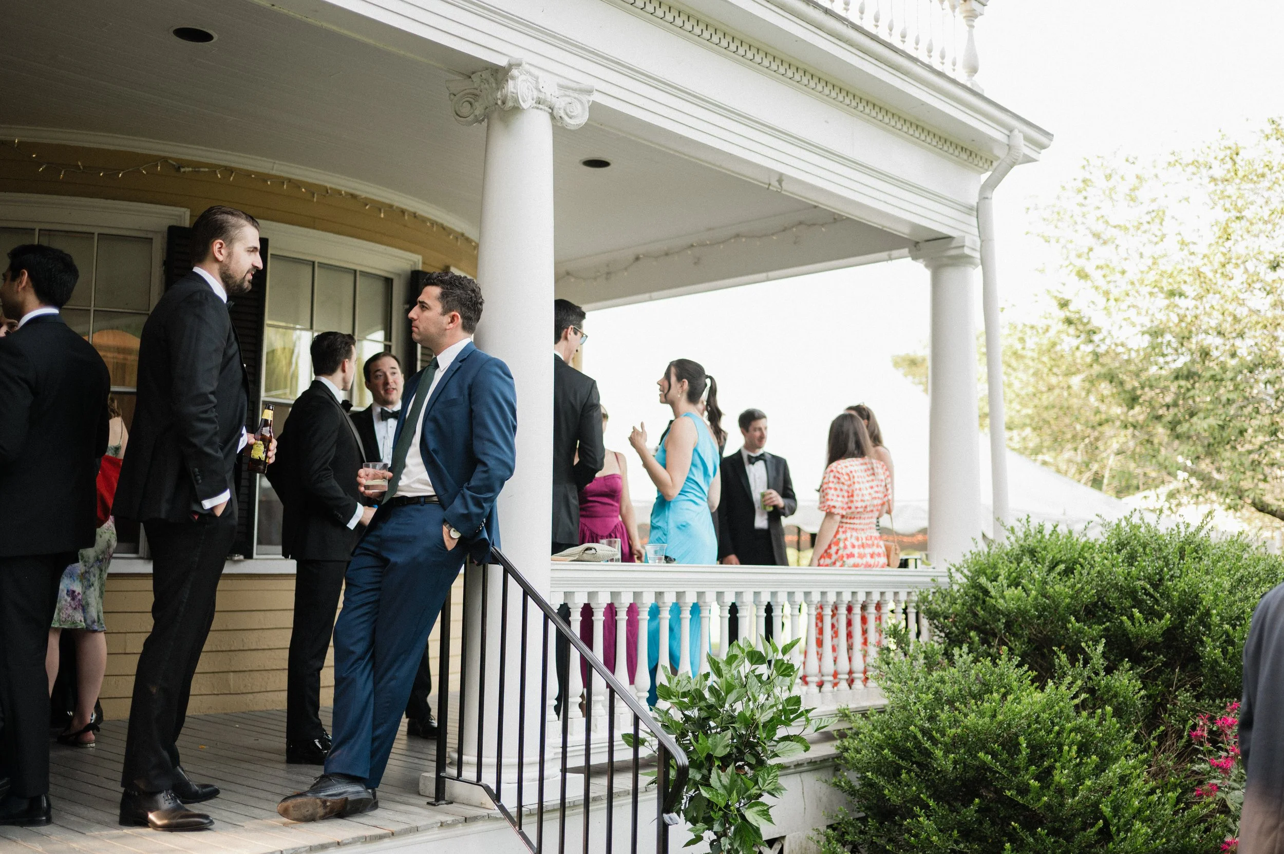 People at a social gathering on a porch, dressed formally, engaging in conversations, with some holding drinks.