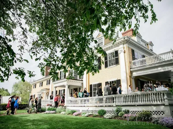People gathered outdoors on a porch and lawn at a large, historic house during daytime.