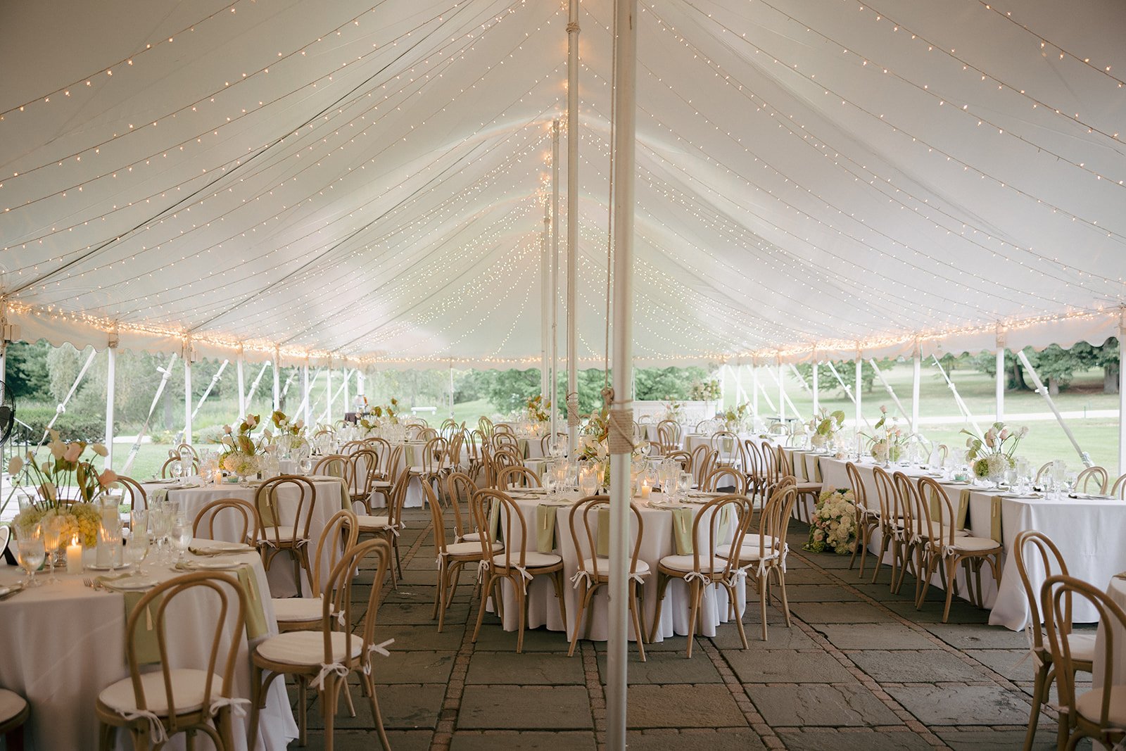Elegant outdoor wedding reception under a large white tent with string lights, round tables covered with white tablecloths, floral centerpieces, and wooden chairs.