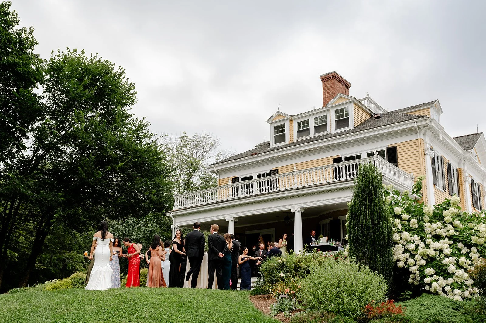 People gathered outside a large, white, Victorian-style house for a social event, with trees, bushes, and flowers in the garden.