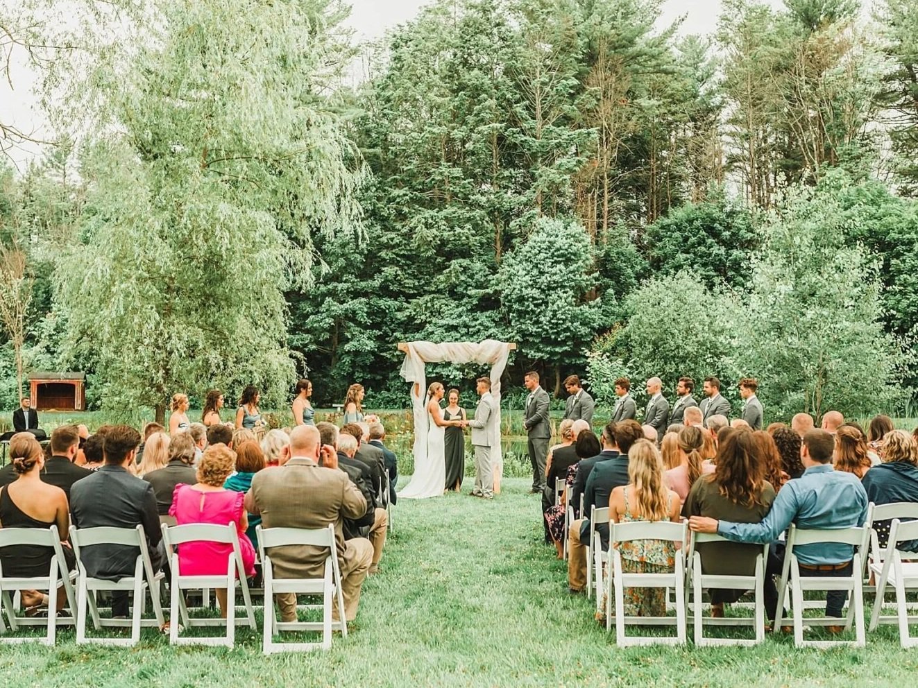Outdoor wedding ceremony with the bride and groom exchanging vows under an arch, surrounded by family and friends seated on white chairs, lush green trees in the background.