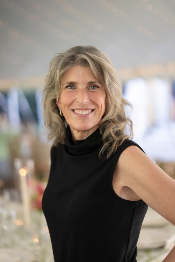A smiling woman with shoulder-length blonde hair wearing a sleeveless black top, standing at an event with blurred lights and decorations in the background.