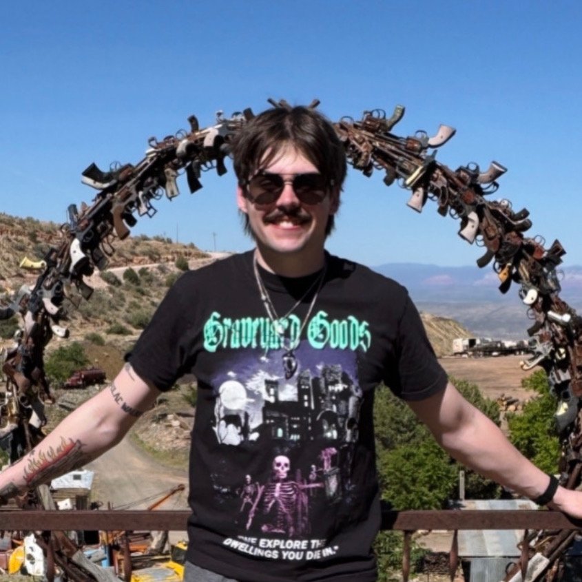 Man with sunglasses and mustache standing outdoors, smiling, with arms outstretched, in front of a sculpture made of various guns and weapons, with a landscape background.