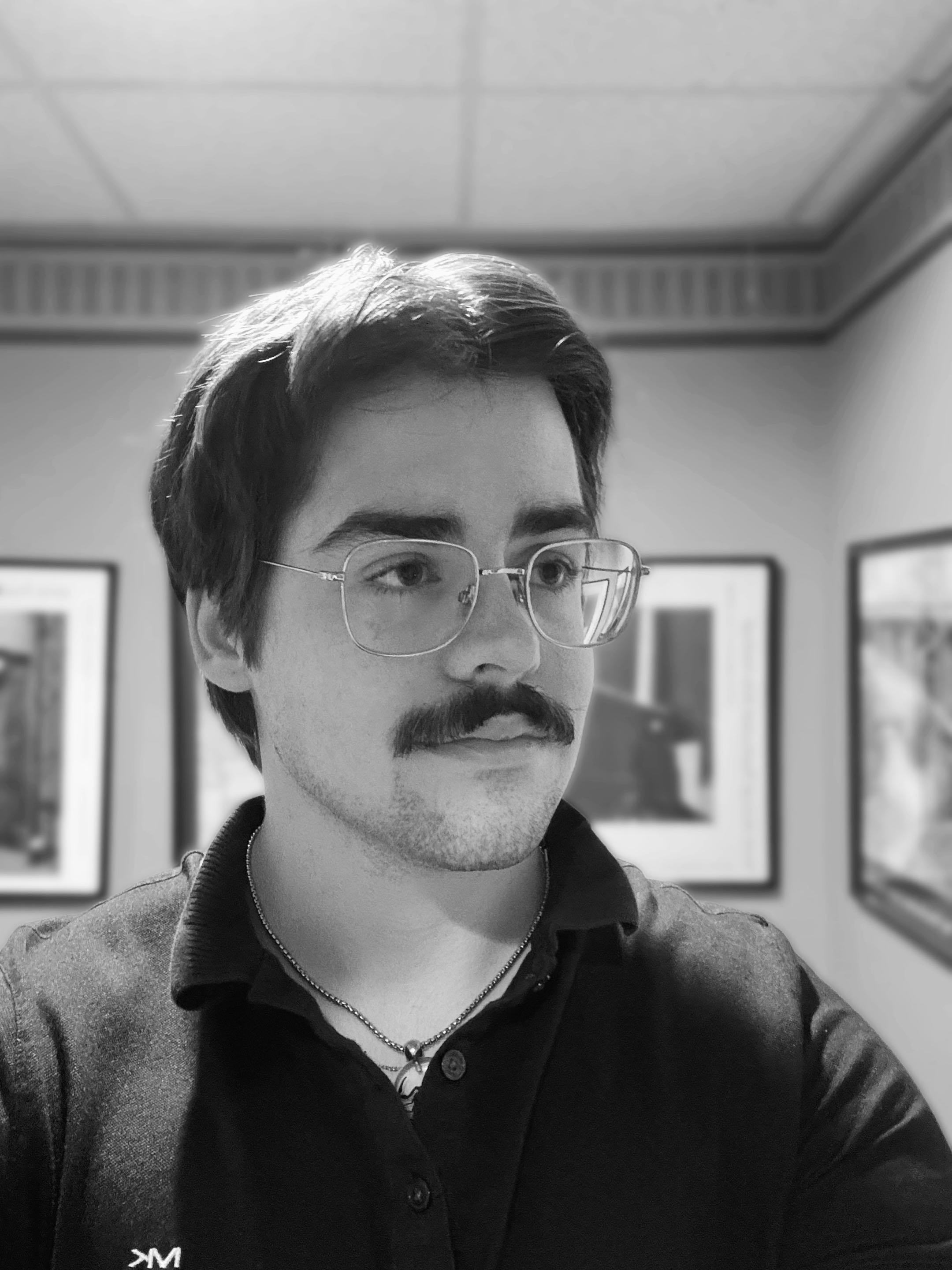 A black and white photo of a person with medium-length hair and glasses, wearing a collared shirt and a silver necklace, looking to the right. There are framed pictures hanging on the wall behind them.