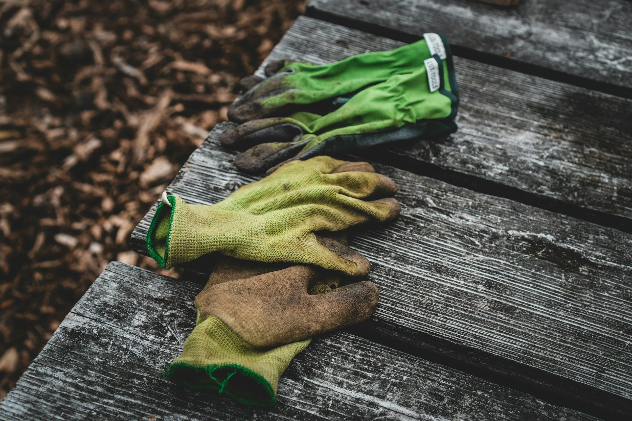 Pair of work gloves, one yellow and one green, resting on a worn wooden bench outdoors.