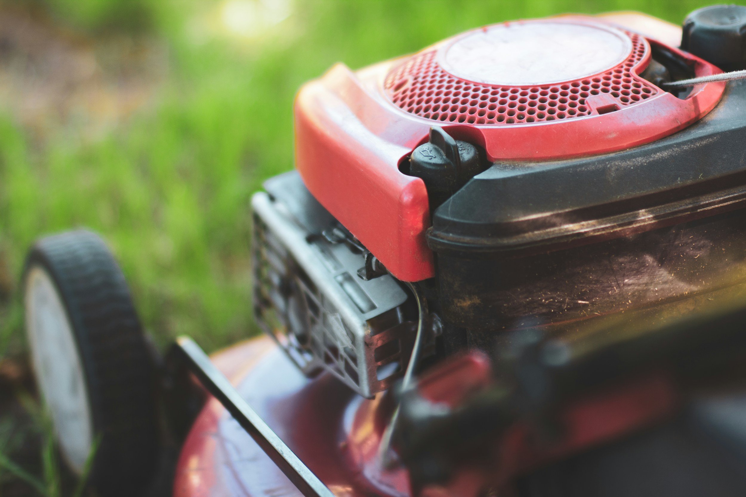 Close-up of a red and black lawn mower with a gray grass cutting blade, set against a blurred green and brown outdoor background.