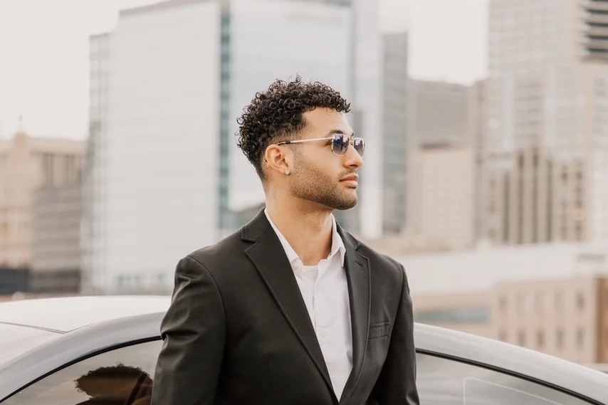 A man in a black suit and white shirt standing outdoors next to a white car in an urban cityscape with tall buildings.