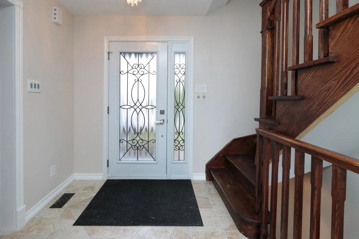 Interior front door entryway with a black doormat, a white door featuring decorative metal grille, a matching side window with metal grille, beige walls, a dark wooden staircase on the right, light-colored tile flooring, and a hanging light fixture.