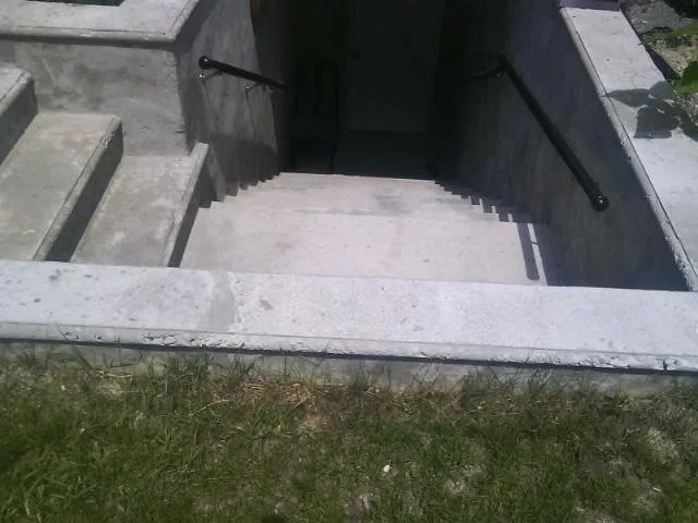Concrete cement outdoor staircase with handrails leading down to a underground area covered by shadow, bordered by grass and concrete walls.