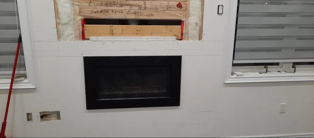 Interior wall insulation and wooden framework in a room, in between two windows with closed white blinds and above a built-in black fireplace, and a red broom resting on the left window.