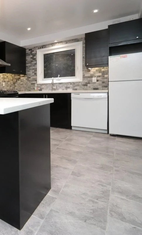 Kitchen with black cabinets, a gray and white brick backsplash, white sparkle marbled countertops, a window above the sink with a thick white frame, a white fridge and dishwasher on the right, tiled grey floors, illuminated by pot lights.