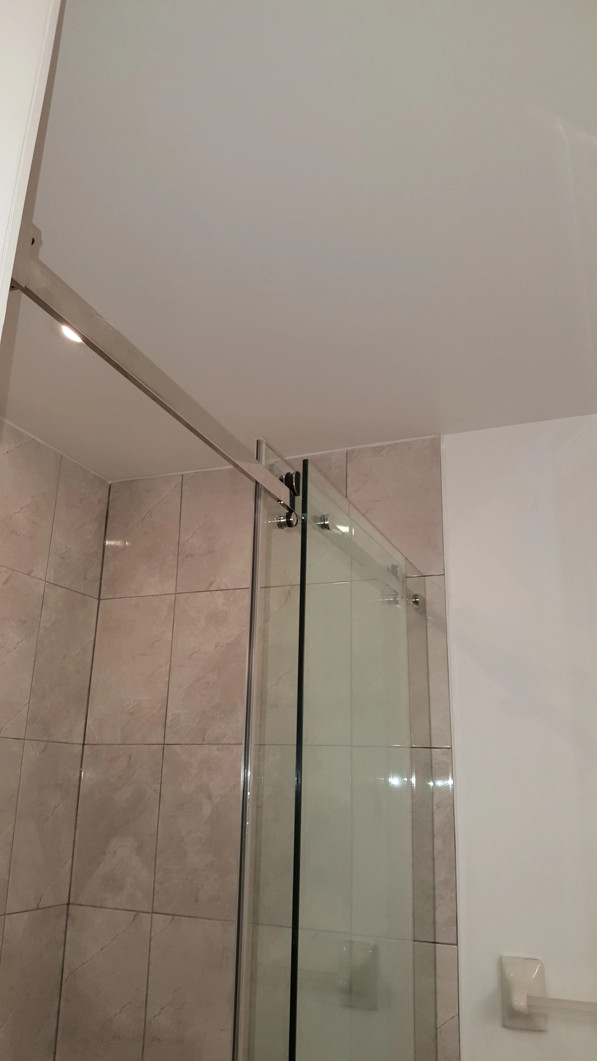View of a bathroom shower area with glass door, beige tiled shower walls, and a white ceiling and white bathroom walls.
