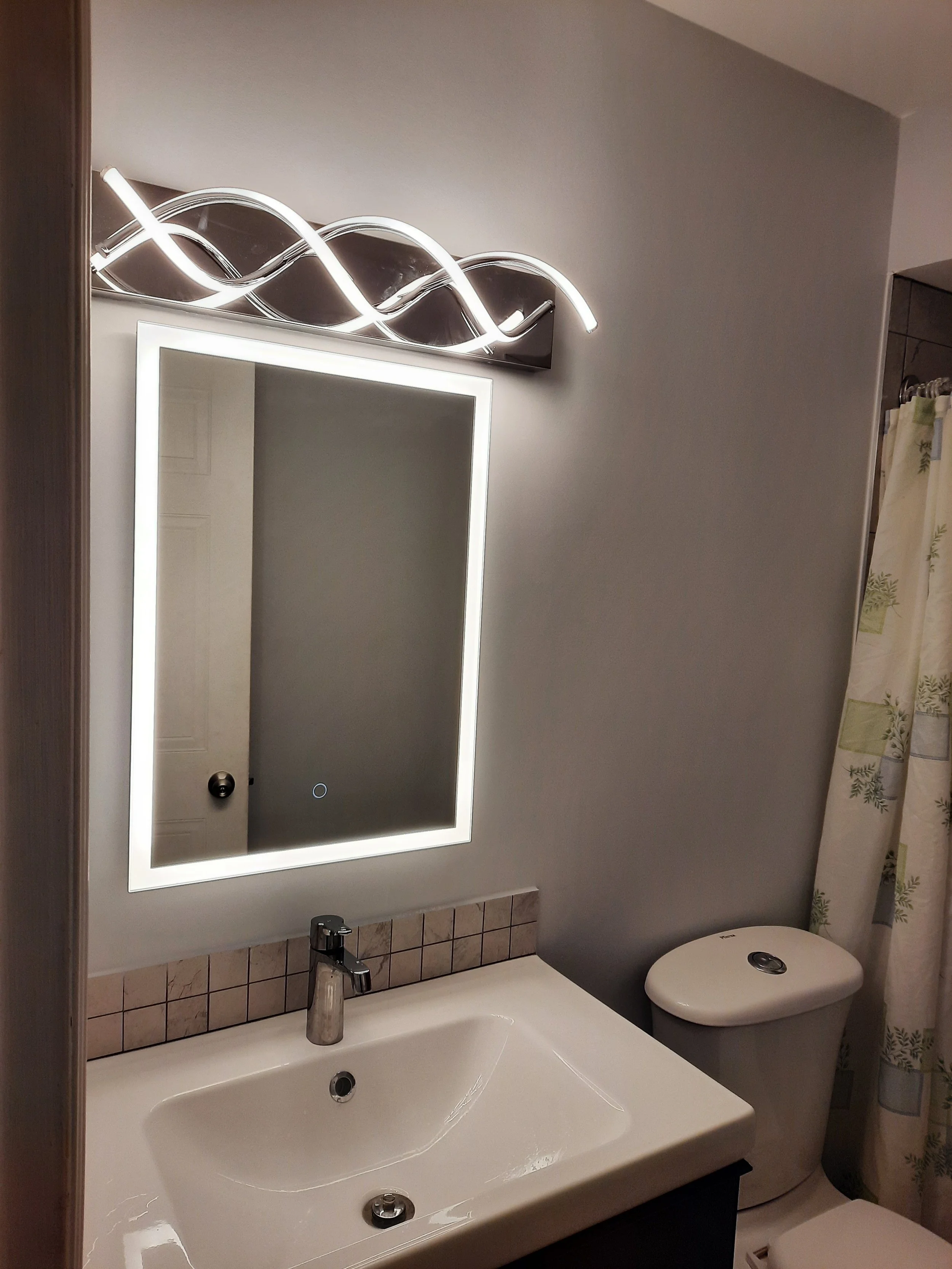 Bathroom with illuminated mirror above sink, toilet, and shower curtain slightly showing with leaf pattern.