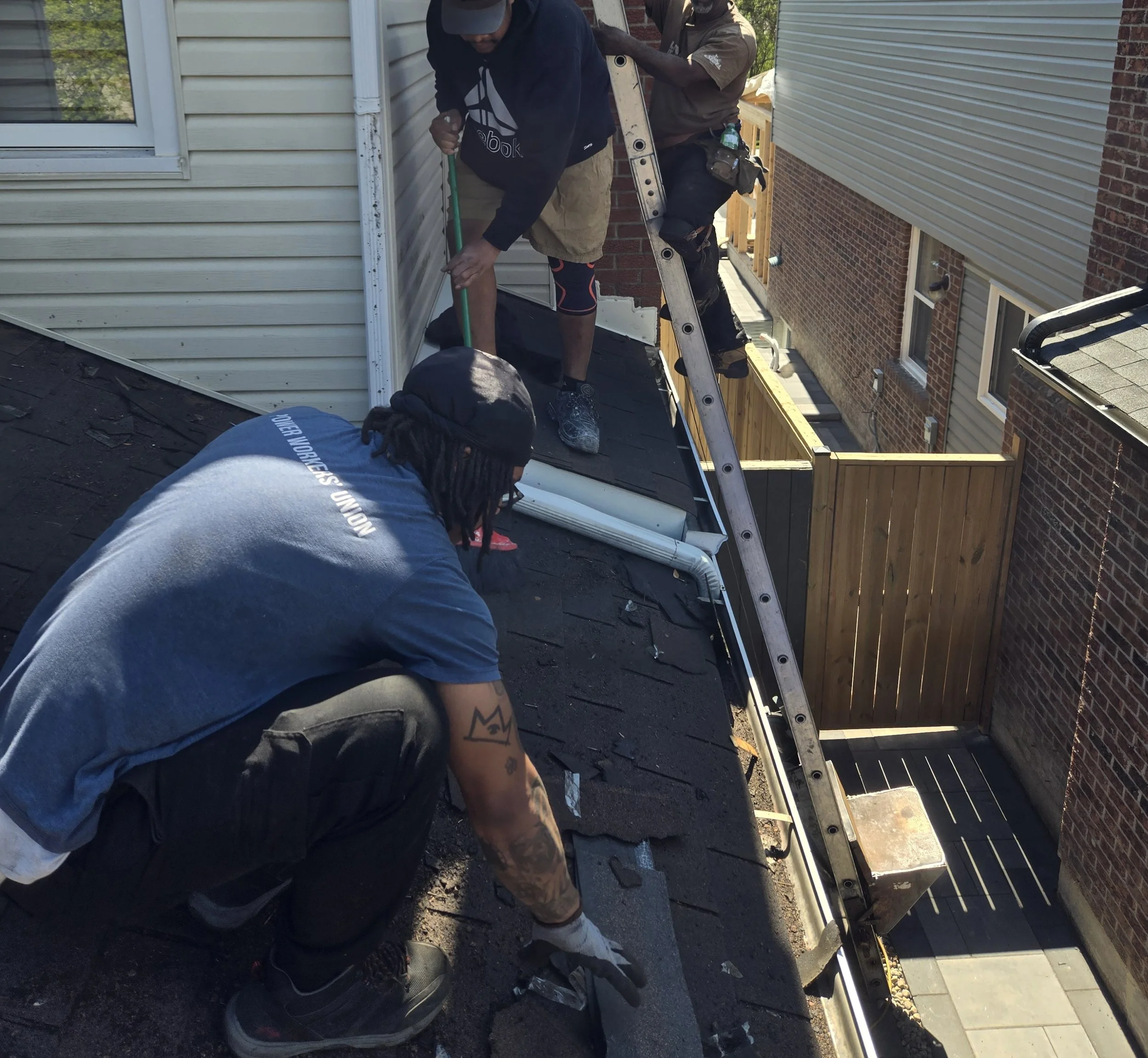 Three men working outside on a house roof, with one man on a ladder, and the other two men working directly on the roof, in the motion of placing and stapling down shingles to the roof.