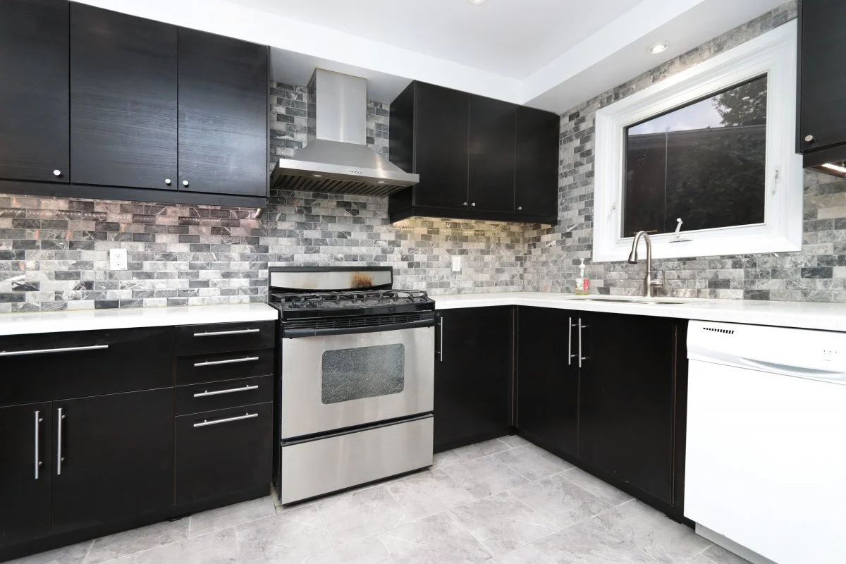 Modern style kitchen with black cabinets, a stainless steel stove and range hood, a gray and white brick backsplash, white sparkle marbled countertops, a window above the sink with a thick white frame, and a white dishwasher.