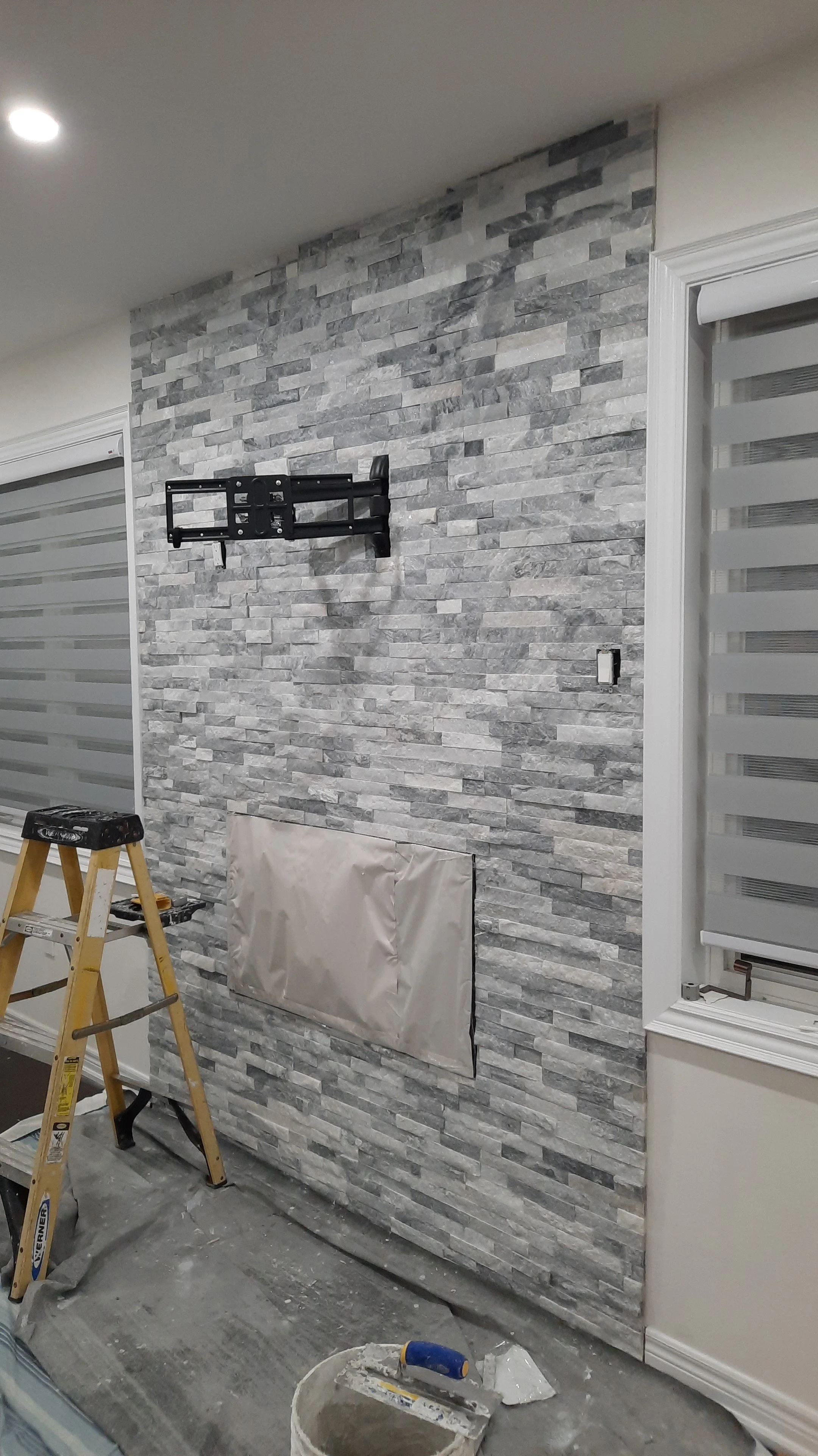 Interior wall with backsplash gray stone tiles, a mounted TV bracket, a ladder, a covered electrical outlet, and construction tools and materials.